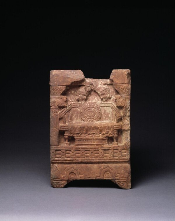 Red sandstone relief of an empty throne with a draped seat and a round wheel-like disc on its backrest, framed by pillars, with two small figures above