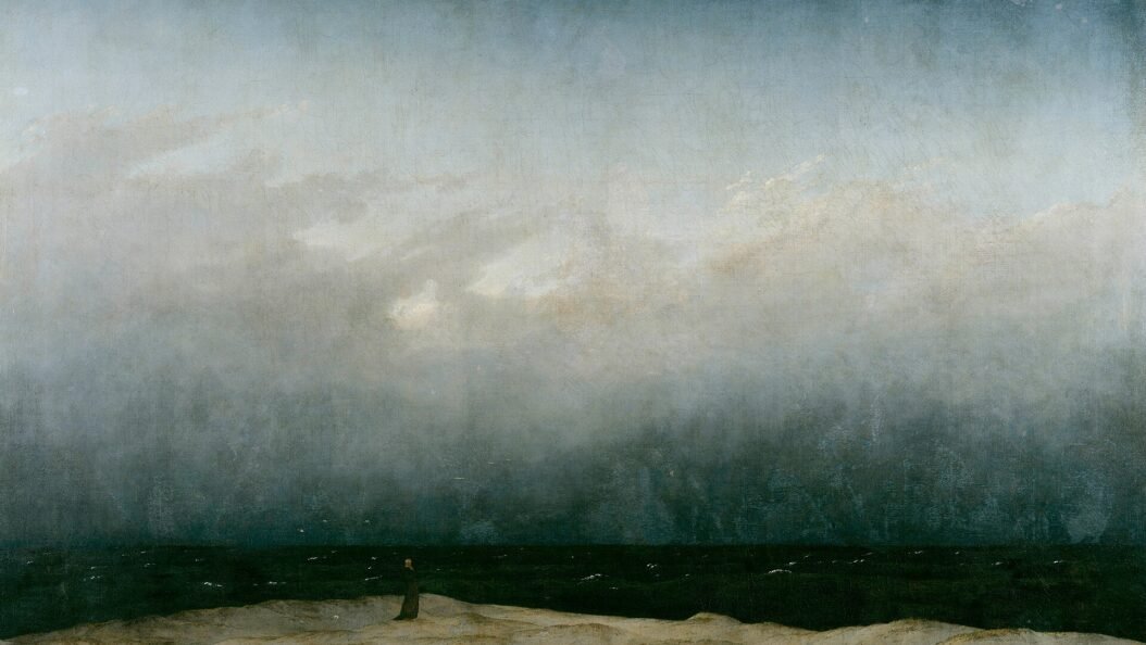 A tiny figure in a dark robe stands alone on a pale strip of dune beside a dark sea, under a vast grey sky that fills most of the painting.