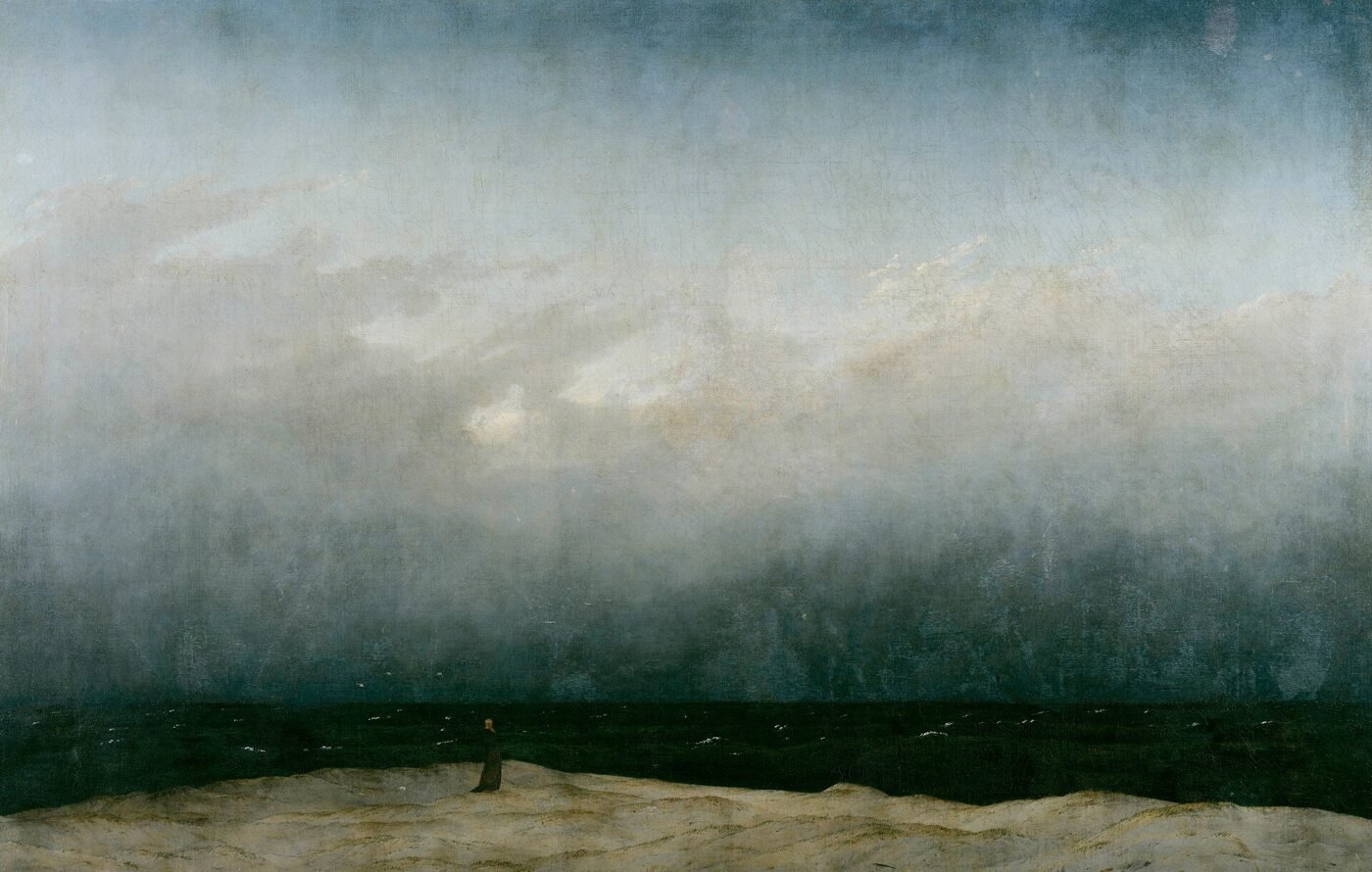 A tiny figure in a dark robe stands alone on a pale strip of dune beside a dark sea, under a vast grey sky that fills most of the painting.